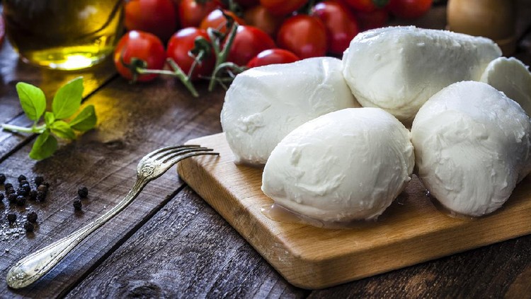 Mozzarella cheese on a cutting board shot on rustic wooden table. On the table all around the board are ingredients for preparing Caprese salad like tomatoes, olive oil, basil, pepper and salt. Low key DSRL studio photo taken with Canon EOS 5D Mk II and Canon EF 100mm f/2.8L Macro IS USM