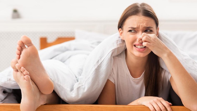 Stinky Feet. Disgusted Girlfriend Pinching Nose Lying Near Boyfriend's Smelly Legs In Bed In Bedroom. Selective Focus