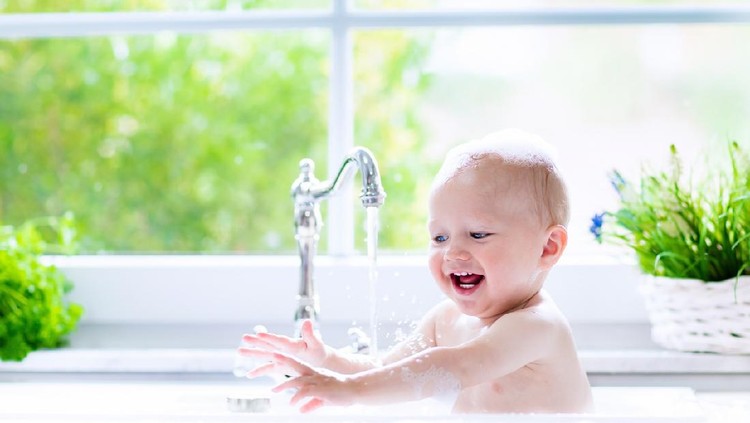 Baby taking bath in kitchen sink. Child playing with foam and soap bubbles in sunny bathroom with window. Little boy bathing. Water fun for kids. Hygiene and skin care for children. Bath room interior