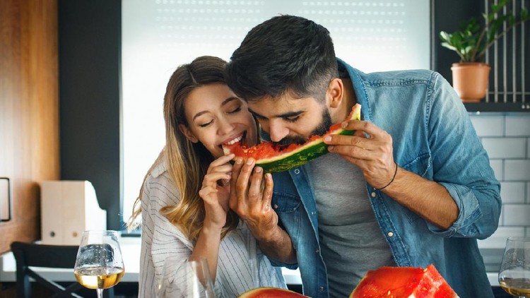 Closeup front view of a mid 20's couple sharing a watermelon in a kitchen. They are biting the same slice and laughing, having some white wine as well