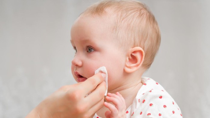 Young mother hand wiping baby girl cheek with white cotton pad. Daily routine. Care about infant clean and soft body skin. Closeup. Side view.