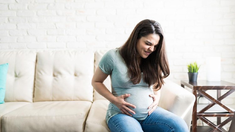 Attractive pregnant female grimacing with hands on baby bump while sitting at home