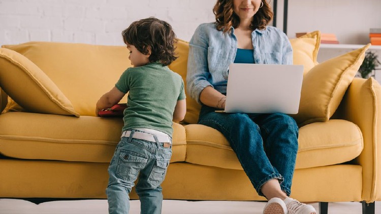 Work From Home kid standing near sofa while mother working with laptop
