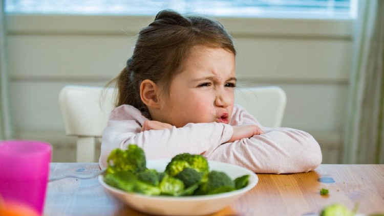Cute girl eating spinach and broccoli at the table. Child doesn't want to eat, refuses eating, making faces. Healthy food concept.