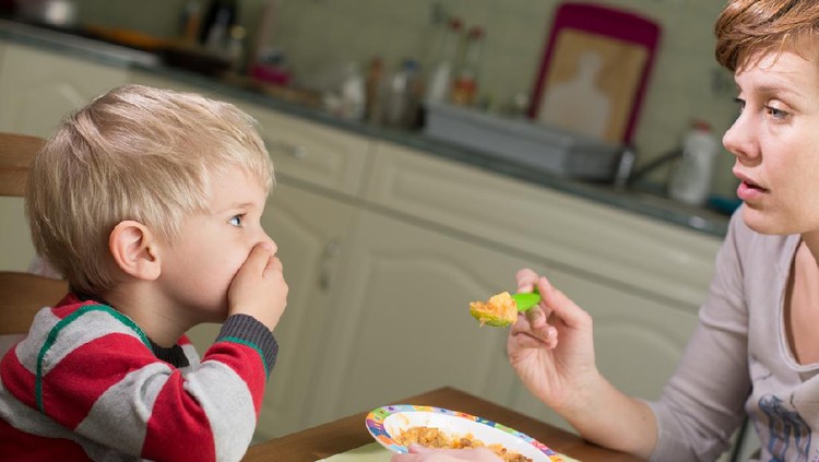 A 3-4 years boy is trying to stop his mother from feeding him. One of the options is to hold a hand on his mouth. Son holds a cool look to his mom, while she is still super-controlling her nerves.