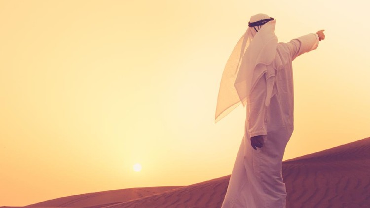 An Arab standing at the sand dunes of Dubai and aiming towards the horizon.
