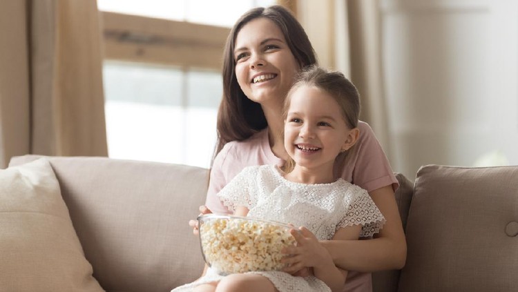 Happy young mom and child girl laughing holding snack popcorn remote control enjoy funny television comedy movie, smiling family mother with kid girl watching fun humor tv show laugh sitting on sofa