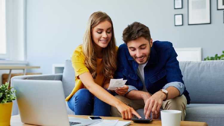 Happy young couple paying bills together and managing budget, sitting on the sofa and using calculator and laptop