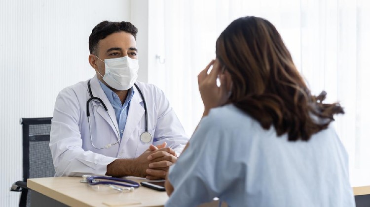 Male doctor wearing face mask discussing with woman patient while sitting on the table in the office at the hospital
