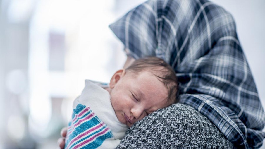 A Muslim woman is laying in a hospital bed. She is holding her newborn baby over her shoulder while he sleeps.