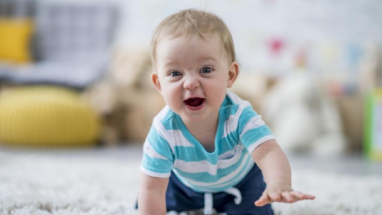 A Caucasian baby boy is crawling towards the camera while laughing. He is giggling and looking at the camera. He is at a daycare center.