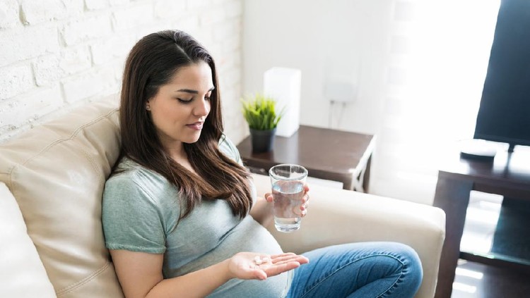 Good looking expectant female looking at pills and holding glass of water in living room