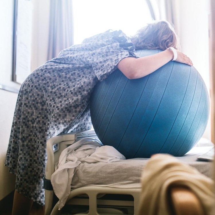 A woman in labor prepares to give birth in a clean white hospital setting. She leans on a birthing or fitness ball to ease the intensity of her contractions.  A depiction of a positive pregnancy and delivery.