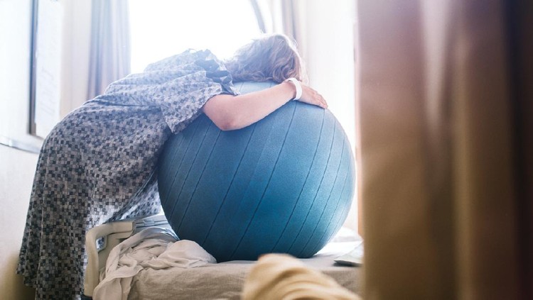 melahirkan A woman in labor prepares to give birth in a clean white hospital setting. She leans on a birthing or fitness ball to ease the intensity of her contractions. A depiction of a positive pregnancy and delivery.