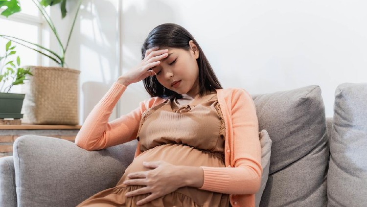 Young pregnant Asian woman has suffered from headaches sitting on the sofa. Pregnancy symptoms