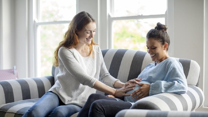 Friend touching pregnant woman's abdomen. Happy females are sitting on striped sofa. They are at home.