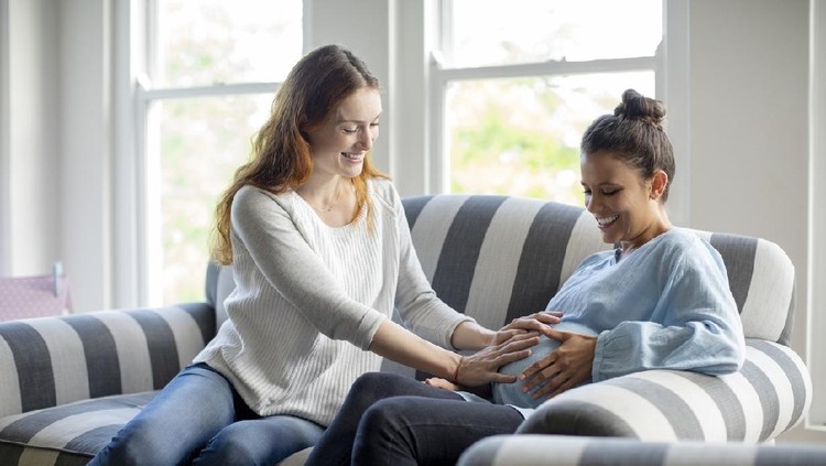 Friend touching pregnant woman's abdomen. Happy females are sitting on striped sofa. They are at home.
