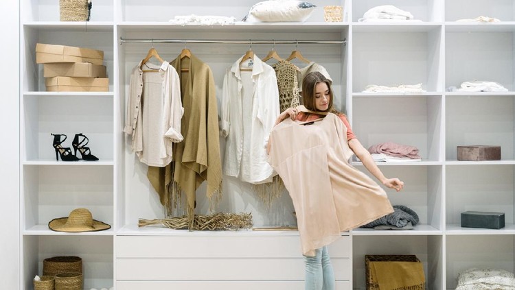 Young adult woman standing near modern wardrobe at home with contemporary interior, choosing trendy clothes, having difficult choice not knowing what to put on, trying on fashionable wear