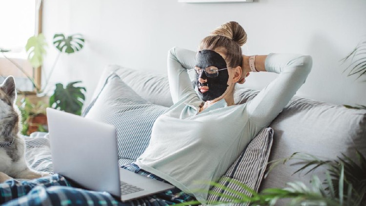 Mature women home in pajama spending cozy morning during isolation period. She wearing sheet mask and have video call, her dog is in bed too