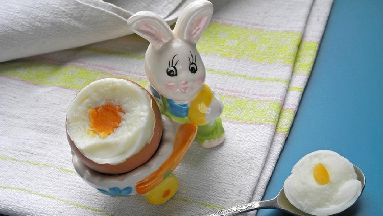 Broken hard boiled egg in decorative eggcup in form of rabbit with wheelbarrow, spoon with piece of egg on linen napkin. Egg white and yolk. Baby and diet food.