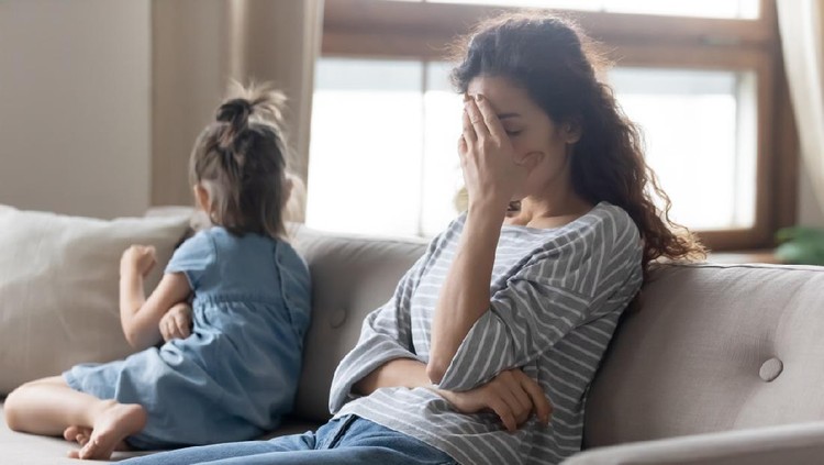 Unhappy young mother touching forehead, feeling tired of bad daughter's behavior at home. Offended little child girl sitting on different side on couch, ignoring sad frustrated mother in living room.