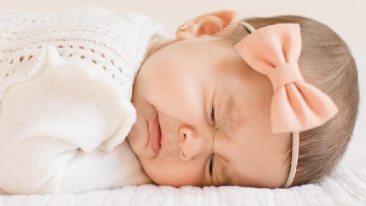 bayi perempuan A 6-month old baby girl wearing a white outfit and a peach-colored bow laying on a white blanket, sneezing.