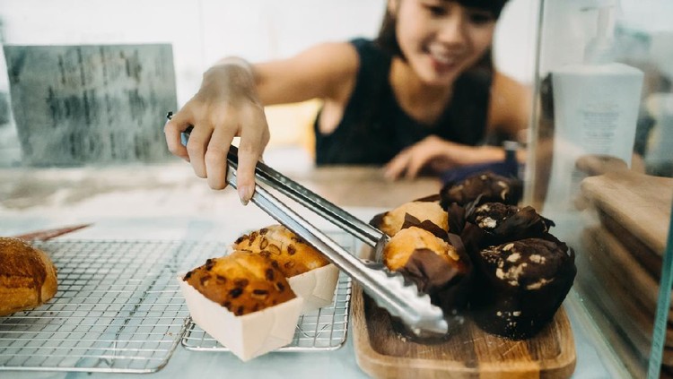 Cheerful young Asian woman working at a bakery and serving fresh bakery from the display cabinet
