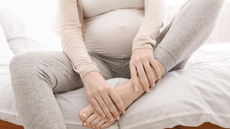 Pregnancy problems. Pregnant woman massaging her swollen foot, sitting on bed