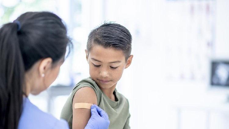 A young mixed-race boy visits a female doctor.  The Doctor is placing a bandage on the boy's arm where he received a vaccine injection.