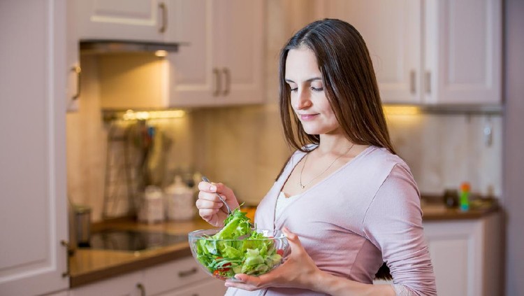 A young beautiful pregnant woman eating a fresh green salad. Healthy nutrition and pregnancy. Pregnant woman's standing in the kitchen with vegetable salad. Healthy lifestyle.