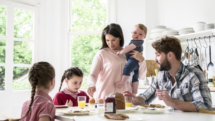 Two girls having breakfast with their dada as their mum carries their younger brother