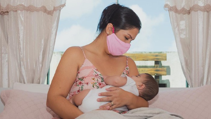 Two people, mother with protective mask breastfeeding her baby son at home, they are at home do to pandemic outbreak quarantine.