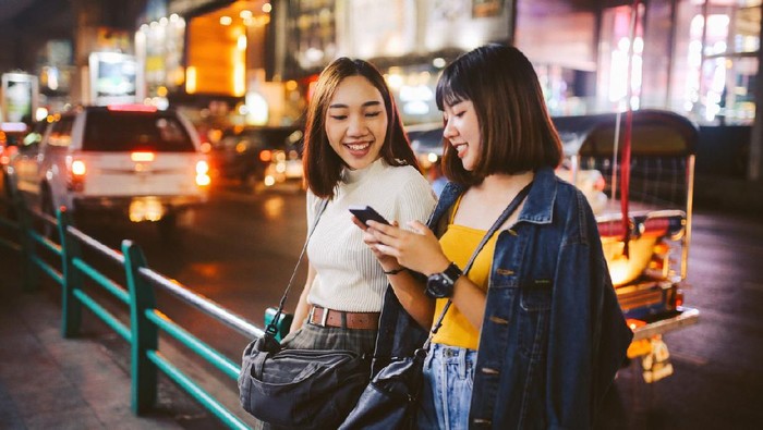 Vintage toned image of two young, Asian women, walking together, chatting, near Siam station on Bangkok Sukhumvit line. They’re wearing casual street style clothing, enjoying an afternoon together, texting on their cellphones.