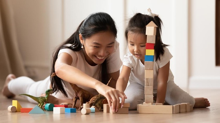 Overjoyed young Asian mom and little biracial daughter lie on warm home floor construct with building bricks together, happy millennial Vietnamese mother have fun play with small girl child