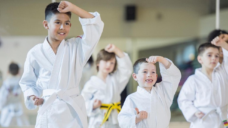 A multi-ethnic group of children are indoors at a Taekwondo academy. They are wearing martial arts clothing. They are practicing their techniques and smiling.