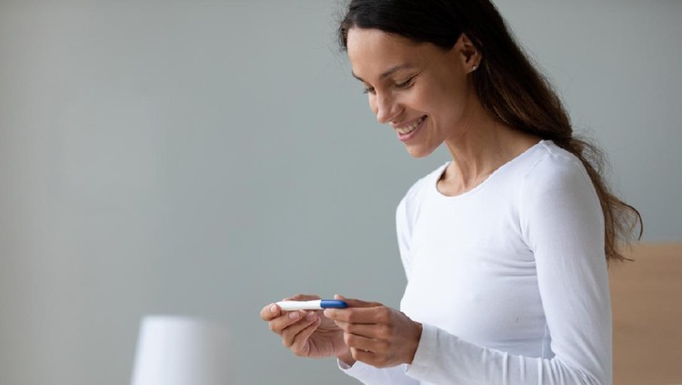 Smiling woman wearing white sleepwear holding pregnancy test, waiting for result close up, sitting in bedroom alone, beautiful young female checking planning pregnancy, fertility maternity concept