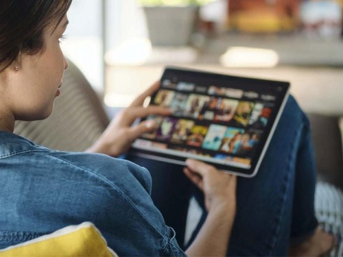 Caucasian woman lying down on sofa in living room at home searching for film or TV series to watch for leisure on her tablet.