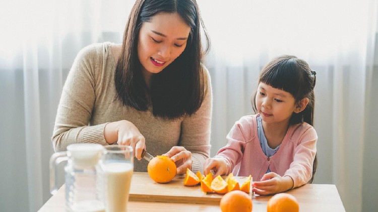 Image of Happy Young Asian mother use kitchen knife cut an orange to pieces with her daughter in living room.