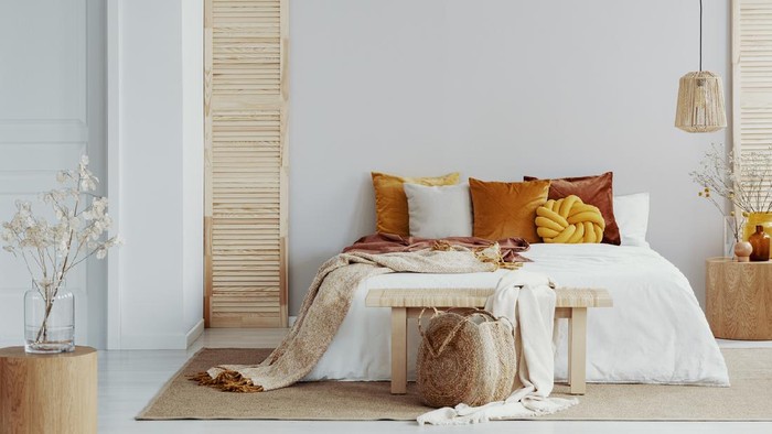 Brown and orange pillows on white bed in natural bedroom interior with wicker lamp and wooden bedside table with vase
