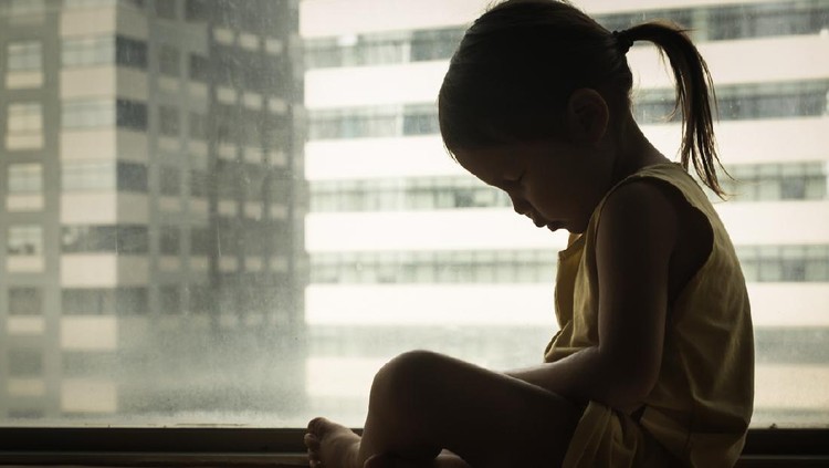 A litte girl sitting next to a window with her head down in sadness. Feeling depressed and hurt.