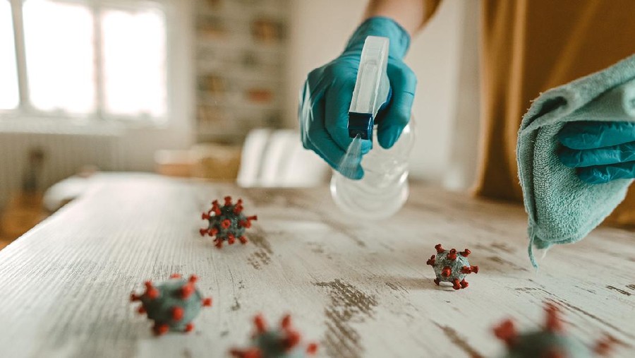 Mature woman cleaning and disinfects table in home office with a disinfectant and a rag. On the table is a model of a corona virus.