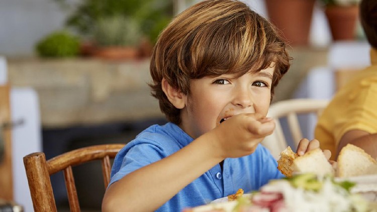 Portrait of boy eating food by table. Male is having lunch at home. He is wearing blue t-shirt.