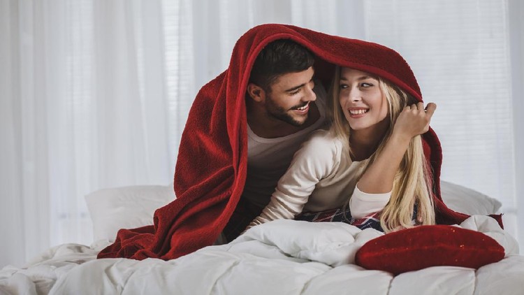 Front view of happy young couple in pajamas decided to have fun by playing under red blanket during long days of quarantine at home.