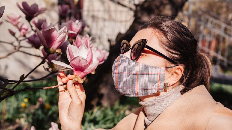 Woman wears reusable mask outdoors during coronavirus covid-19 pandemic. Girl smells magnolia spring flowers. Stay safe, positive. Spring fashion