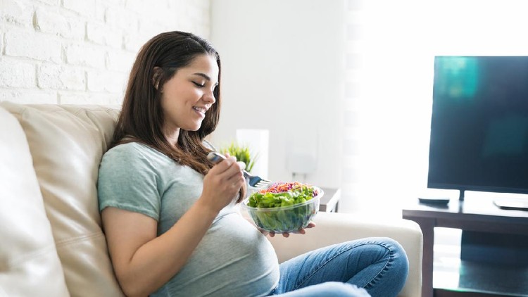 Beautiful expectant woman eating fresh healthy salad from bowl while sitting at home