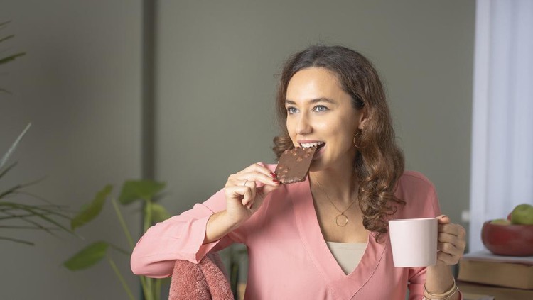 Freelancer young woman having a little coffee break eats chocolate. Comfortable and cheerful work place in her room.