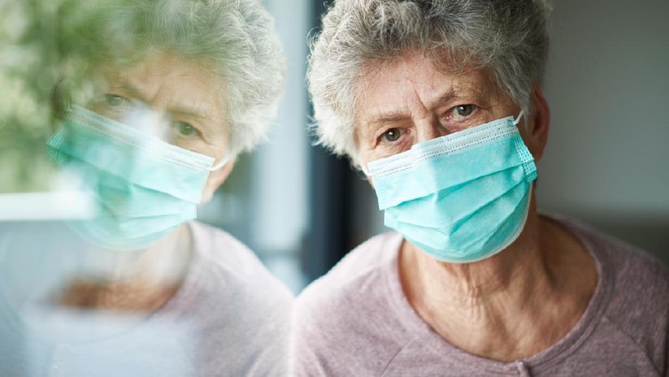 a old woman or grandma is wearing a respirator or surgical mask and is looking out the window while she is in quarantine because of the corona virus
