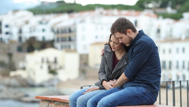 Sad woman and man comforting her on a ledge with a town in the background