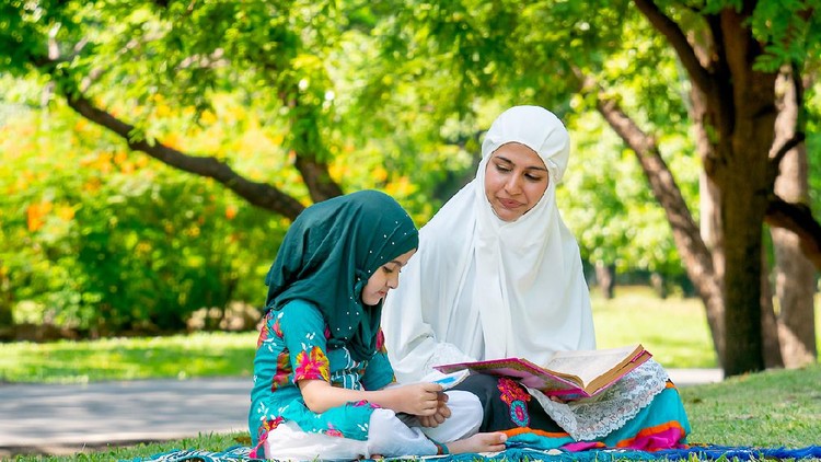 mendongeng Muslim mother teach her daughter to read religion textbook for understanding the way of good life. They stay in the green garden during day time.
