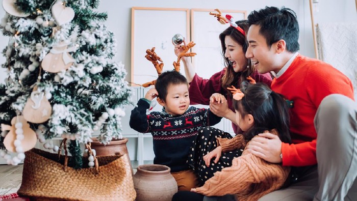 Christmas lifestyle theme. Happy Asian family decorating Christmas tree together in the living room at home. They are putting on various baubles and ornaments and enjoying their holiday